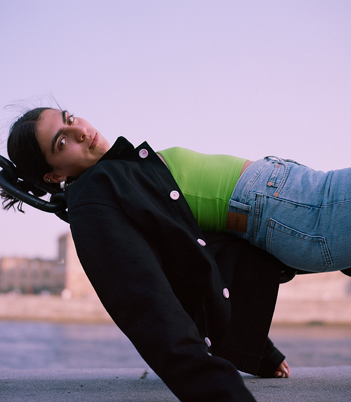 Girl wearing black jacket over a greeen tank and blue denim in front of a natural unset sky